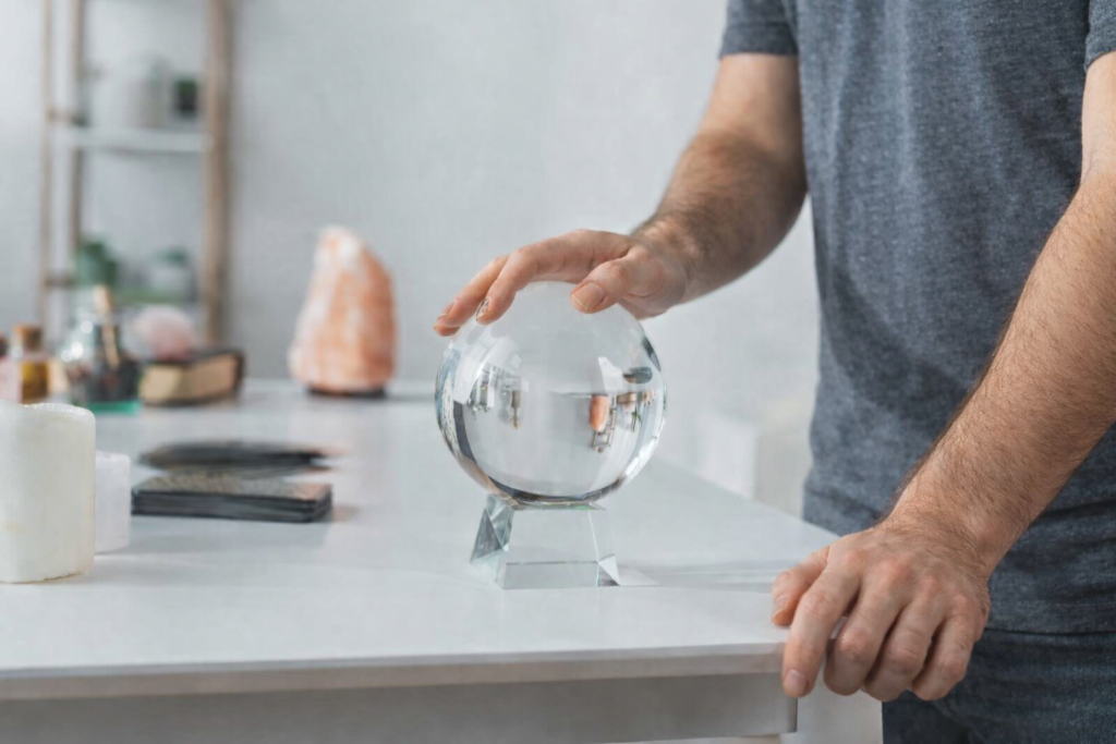 Mãos masculinas tocando uma bola de cristal sobre mesa branca, em ambiente terapêutico minimalista com elementos espirituais ao fundo.